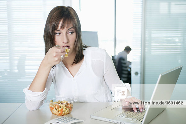 Businesswoman eating pasta and using a laptop