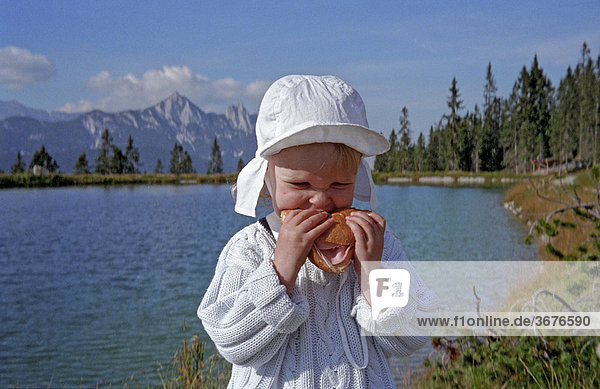 Kleines Mädchen an einem Bergsee beisst herzhaft in eine Wurstsemmel Österreich