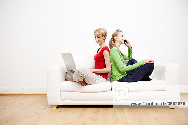 Two women using laptop and phone on sofa