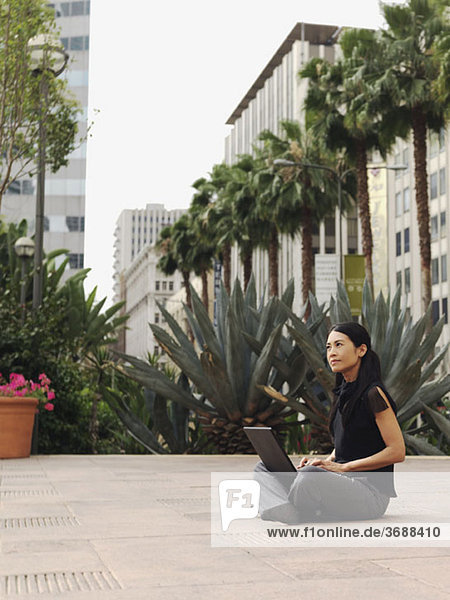 Eine Geschäftsfrau sitzt im Schneidersitz im Hof eines Bürokomplexes  mit einem Laptop.