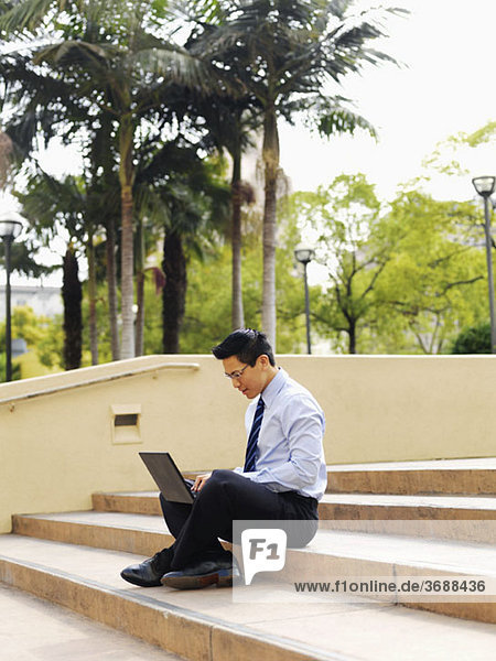Ein Geschäftsmann  der im Schneidersitz auf einer Treppe sitzt und einen Laptop benutzt.