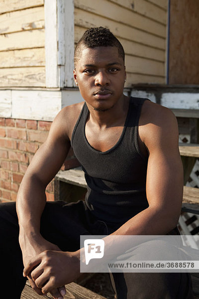 Porträt eines jungen Mannes  der vor einem Haus sitzt.