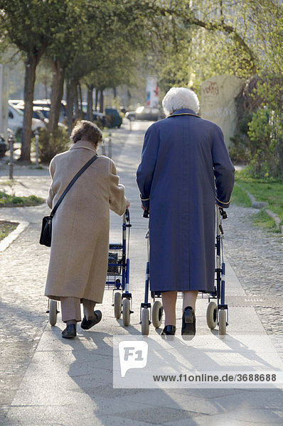 Two women using walkers to walk together on a sidewalk  rear view
