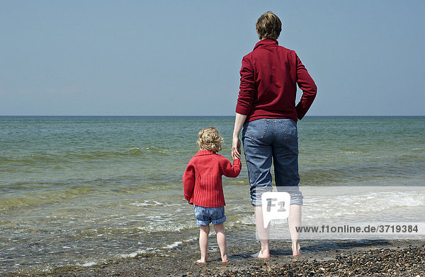 Junge Frau steht mit ihrer kleinen Tochter am Strand und schaut aufs Meer