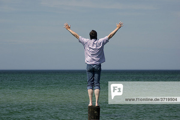 Mann steht auf einem Pfahl in der Ostsee und streckt die Arme in die Luft