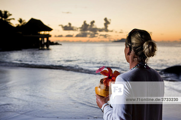 Eine Frau in einer weißen Tunika hält eine dekorierte Kokosnuss in der Hand und schaut auf das Meer  hinten das Sunset Beach Resort  Insel Mahe  Seychellen  Indischer Ozean  Afrika