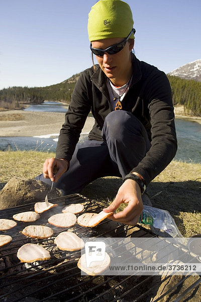 Young woman cooking on a camp fire  roasting ham  barbecue  camping  Takhini River behind  Yukon Territory  Canada  North America