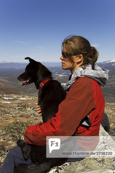 Hiker resting  sitting on a rock  young woman with dog  sled dog  Alaskan husky  mountains behind  Kusawa Ridge  Yukon Territory  Canada  North America