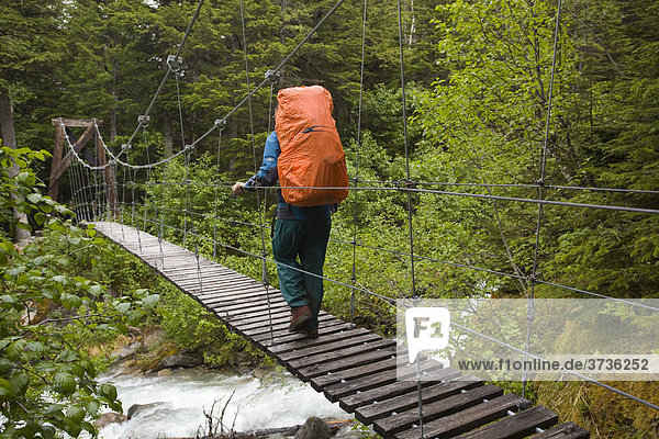 Female hiker with backpack crossing suspension bridge over Taja River  near historic Canyon City  Pacific Northwest Coastal Rain Forest  Chilkoot Trail  Chilkoot Pass  Alaska  USA