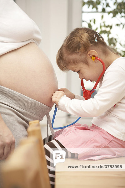 Vierjährige Tochter hört mit Stethoskop den Schwangerschaftbauch der Mutter ab