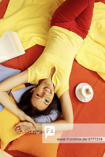 Junge Frau räkelt sich mit Milchkaffee und Buch auf bunten Kissen