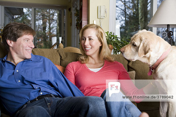 Ein lächelndes Paar sitzt mit seinem Hund auf einer Couch.