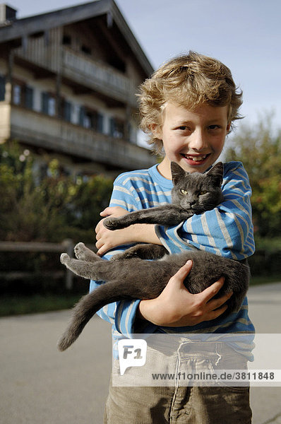 Neun Jahre alter Junge hält Katze. Bayerischer Bauernhof im Hintergrund