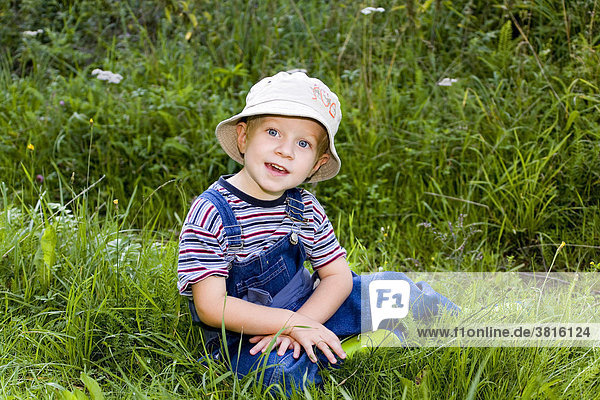 Ein drei Jahre alter Bube mit Schlapphut sitzt im hohen Gras