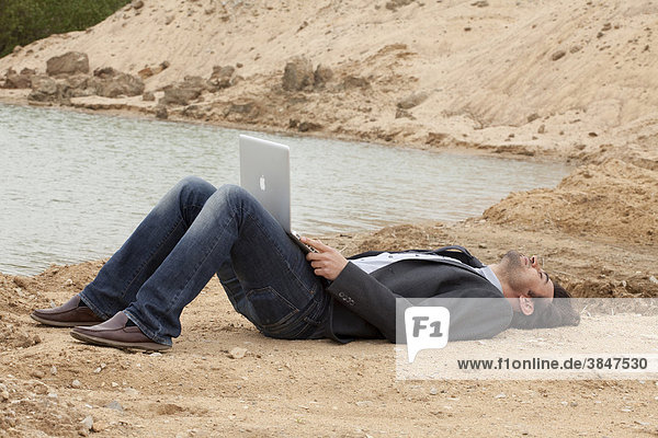 Young man with laptop lying on a lake's beach thinking