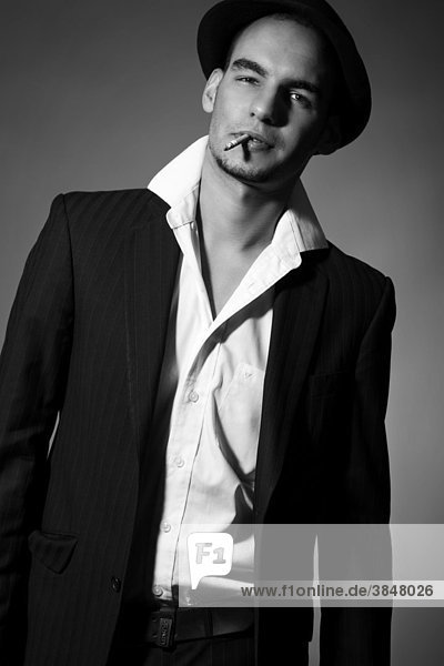 Junger Mann mit Anzug  Hemd  Krawatte und Hut  mit einer Zigarette im Mund und coolem Blick