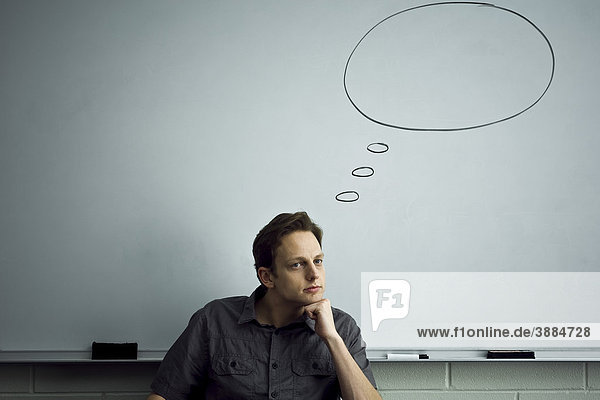 Teacher sitting in classroom contemplatively looking away  empty word bubble above his head