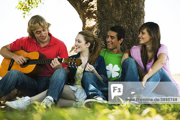 Junger Mann sitzt mit Freunden unter einem Baum und spielt Gitarre.