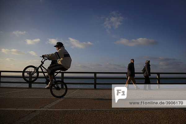 Ein älteres Paar geht an einer Meerpromenade spazieren während ein Junge auf seinem Mountainbike Künststücke vollführt  Alabasterküste  Normandie  Frankreich  Europa