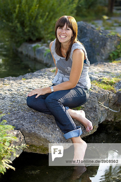 Eine Frau sitzt auf einem Stein am Wasser  jung  lächelnd
