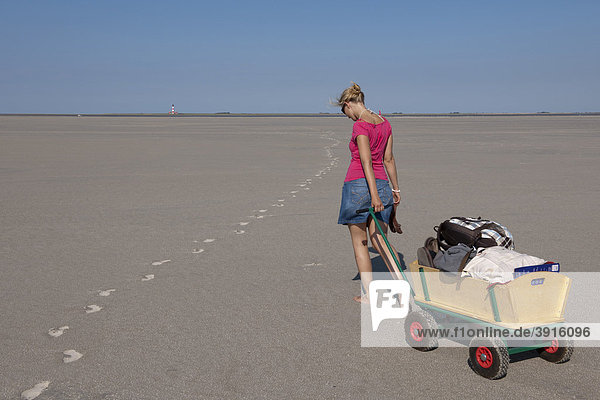 Junge Frau  20-25 Jahre  zieht ihren Bollerwagen mit Strandgepäck über den Sandstrand von St Peter Ording  Nordsee  Nordfriesland  Schleswig-Holstein  Norddeutschland  Deutschland  Europa