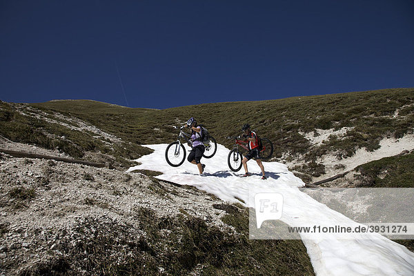 Mountainbike-Fahrerin und Fahrer tragen Räder über Schneefeld auf dem Trail von der Kreuzjochscharte zum Ju dles Cacagnares  Naturpark Fanes-Sennes-Prags  Trentino  Südtirol  Italien  Europa