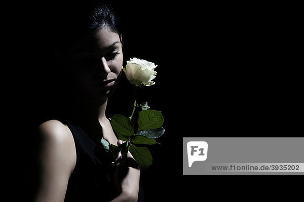 Junge dunkelhaarige Frau sieht traurig zu Boden und hält eine weiße Rose in der Hand