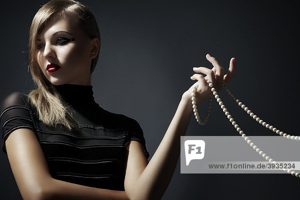 Junge blonde Frau im schwarzen Kleid hält eine Perlenkette in ihrer Hand  Fashion