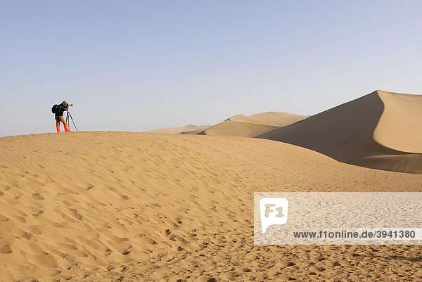 Photographer using a tripod and wearing red boots while standing on a sand hill in front of the sand dunes of the Gobi Desert and Mount Mingshan near Dunhuang  Silk Road  Gansu  China  Asia