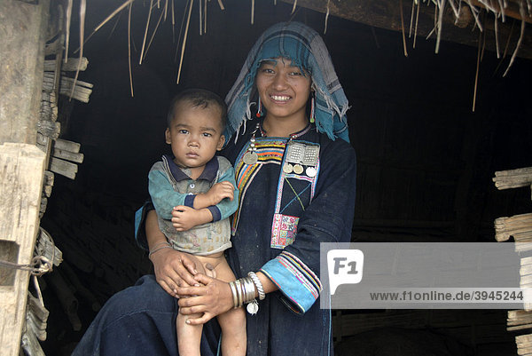 Armut  junge Frau der Akha Nuqui Ethnie  Lächeln  Kleinkind auf dem Schoß  Tracht  traditionelle Kleidung  Farbe Indigo  Kopfbedeckung Tuch  vor Hütte  Dorf Ban Phou Yot  Provinz und Distrikt Phongsali  Laos  Südostasien  Asien