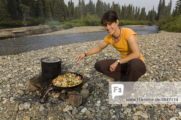 Young woman cooking  frying chicken stir fry on a camp fire  pot  pan  spatula  gravel bar  upper Liard River  Yukon Territory  Canada