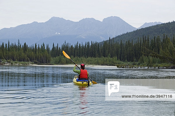 Eine Frau paddelt in einem Kajak auf dem oberen Liard River  dahinter die Pelly Mountains  Yukon Territory  Kanada