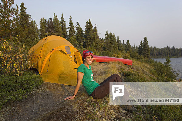 Young woman sitting  enjoying view  relaxing  tent and canoe behind  Caribou Lakes  upper Liard River  Yukon Territory  Canada