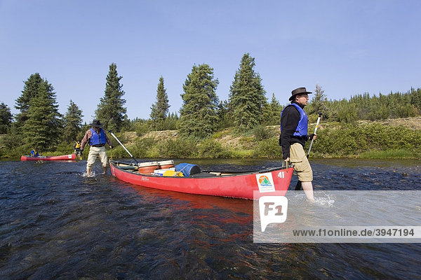 Zwei Männer waten barfuß mit einem Kanu durch den seichten Caribou Creek  oberer Liard River  Yukon Territory  Kanada