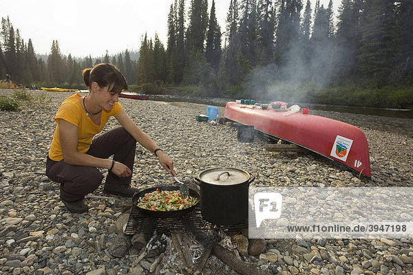 Junge Frau brät Hühnchen in einer Pfanne auf dem Lagerfeuer  dahinter ein Kanu  das als Tisch verwendet wird  Kiesbank  oberer Liard River  Yukon Territory  Kanada
