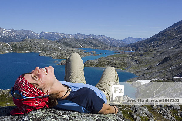 Young woman laughing  lying on a rock  relaxing  hiker enjoying the panorama on summit of historic Chilkoot Pass  Chilkoot Trail  Crater Lake behind  alpine tundra  Yukon Territory  British Columbia  B. C.  Canada