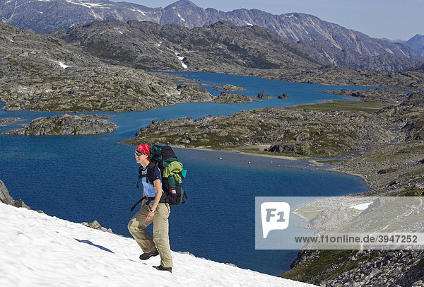 Young woman hiking  backpacking  hiker with backpack  snow field  descending towards summit of historic Chilkoot Pass  Chilkoot Trail  Crater Lake behind  alpine tundra  Yukon Territory  British Columbia  B. C.  Canada