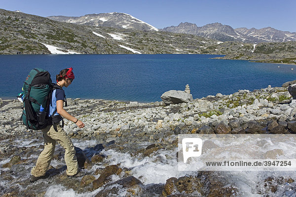 Young woman crossing a creek  hiking  backpacking  hiker with backpack  historic Chilkoot Pass  Chilkoot Trail  Crater Lake behind  alpine tundra  Yukon Territory  British Columbia  B. C.  Canada