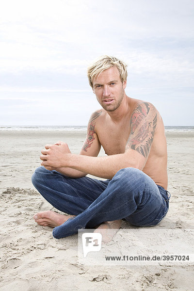 Junger Mann sitzt mit freiem Oberkörper am Strand