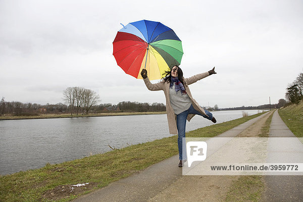 Junge Frau  30-35 Jahre  mit buntem Regenschirm  macht Luftsprünge am Nord-Ostsee-Kanal  Schleswig-Holstein  Deutschland  Europa