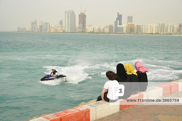 Verschleierte Frauen beobachten Wasserscooter-Fahrer vor der Skyline von Abu Dhabi  Vereinigte Arabische Emirate  Arabien  Naher Osten  Orient