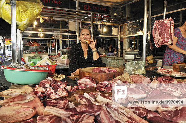 Marktfrau an einem Marktstand mit verschiedenen Fleischstücken  verspeist genüsslich ein Stück Brot  Waren hängen auf Metallhaken in der Luft  Fleischmarkt  Vinh Long  Mekongdelta  Vietnam  Asien