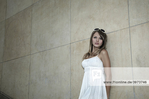 Portrait of a young dark blonde woman in a white dress and sunglasses in front of a marble wall