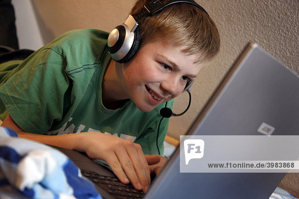 Junge  13 Jahre alt  arbeitet mit seinem Computer zuhause in seinem Kinderzimmer auf dem Bett  surft im Internet auf einer Chat-Seite  spricht mit anderen Chat-Teilnehmern über sein Headset