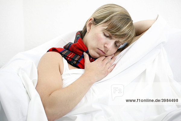 Junge Frau  liegt mit einem Schal um den Hals im Bett  hat eine Erkältung  Halsschmerzen