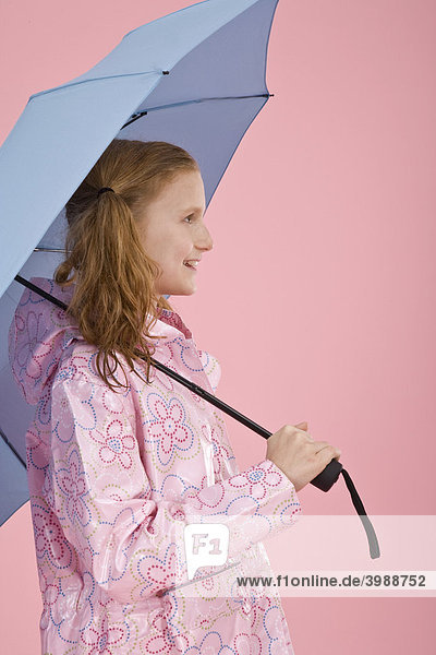Rothaariges Mädchen mit hellblauem Regenschirm und Regenmantel vor Rosa