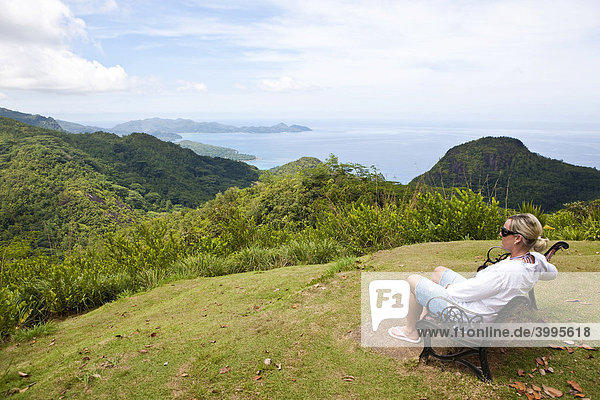 Frau sitzt auf einer Bank im Nationalpark Morone  hinten der Strand von Grand Anse Insel Mahe  Seychellen  Indischer Ozean  Afrika