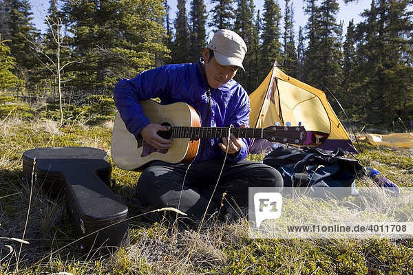Asiat sitzt im Gras und spielt Gitarre  dahinter ein Zelt  Camping  Yukon Territory  Kanada  Nordamerika
