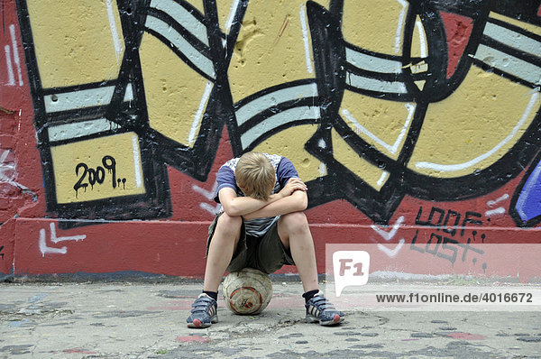 Enttäuschter und einsamer Junge mit seinem Fußball vor einer Graffitiwand  Bolzplatz in Köln  Nordrhein-Westfalen  Deutschland  Europa