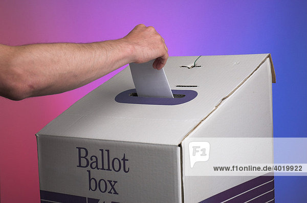 Hand wirft Stimmzettel in Wahlurne vor farbiger Fläche mit gegensätzlichen Farben des politischen Spektrums